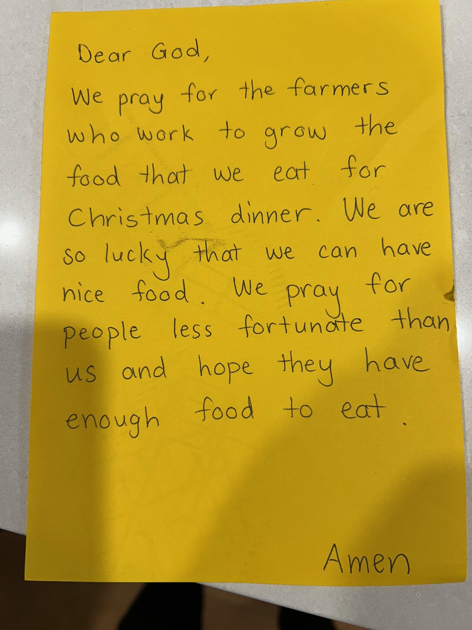 A yellow piece of paper with the following handwriting: Dear God, We pray for the farmers who work to grow the food that we eat for Christmas dinner. We are so lucky that we can have nice food. We pray for people less fortunate than us and hope they have enough food to eat. Amen