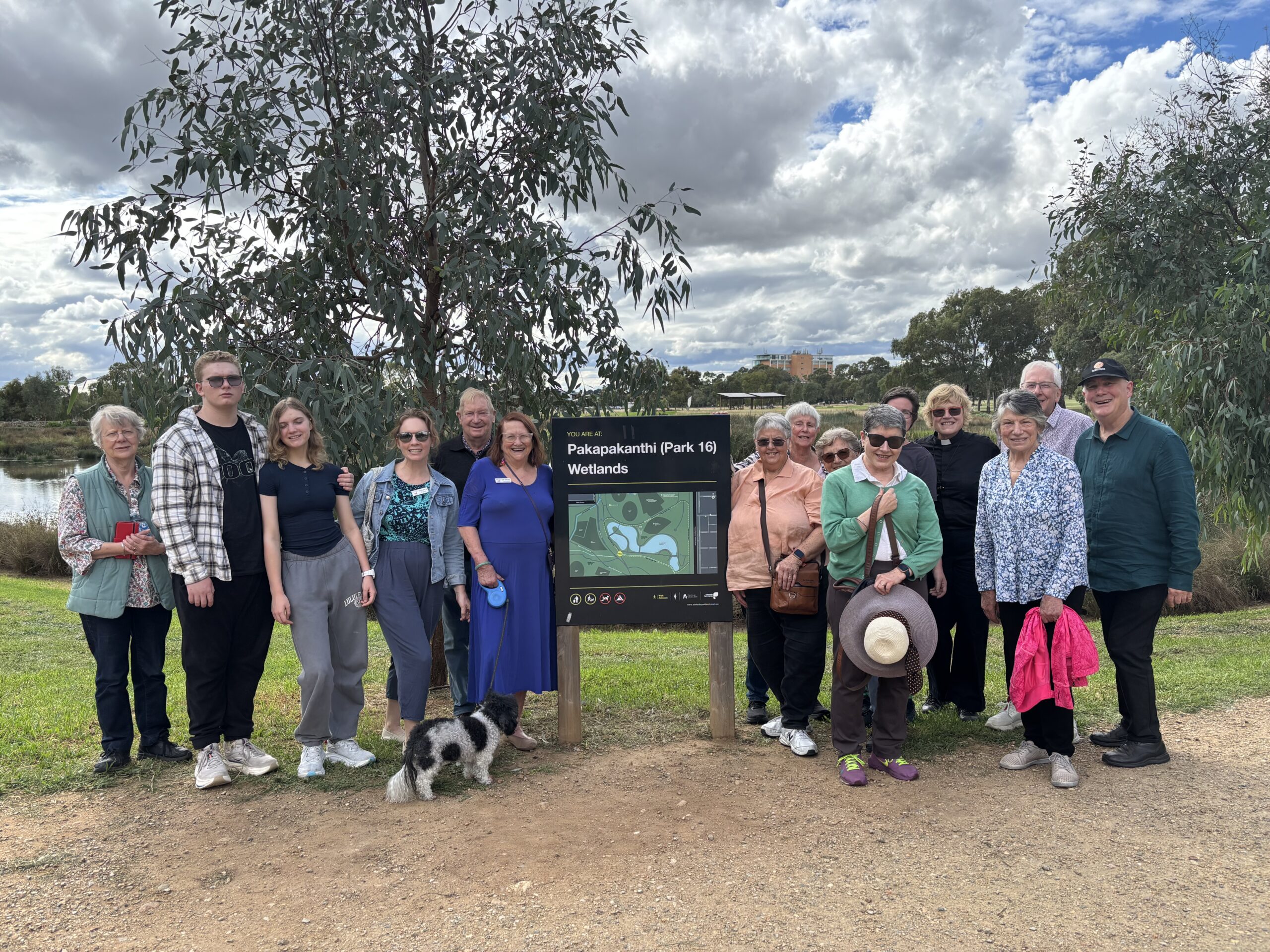 Group of 15 smiling people standing around a sign in a leafy wetland park.