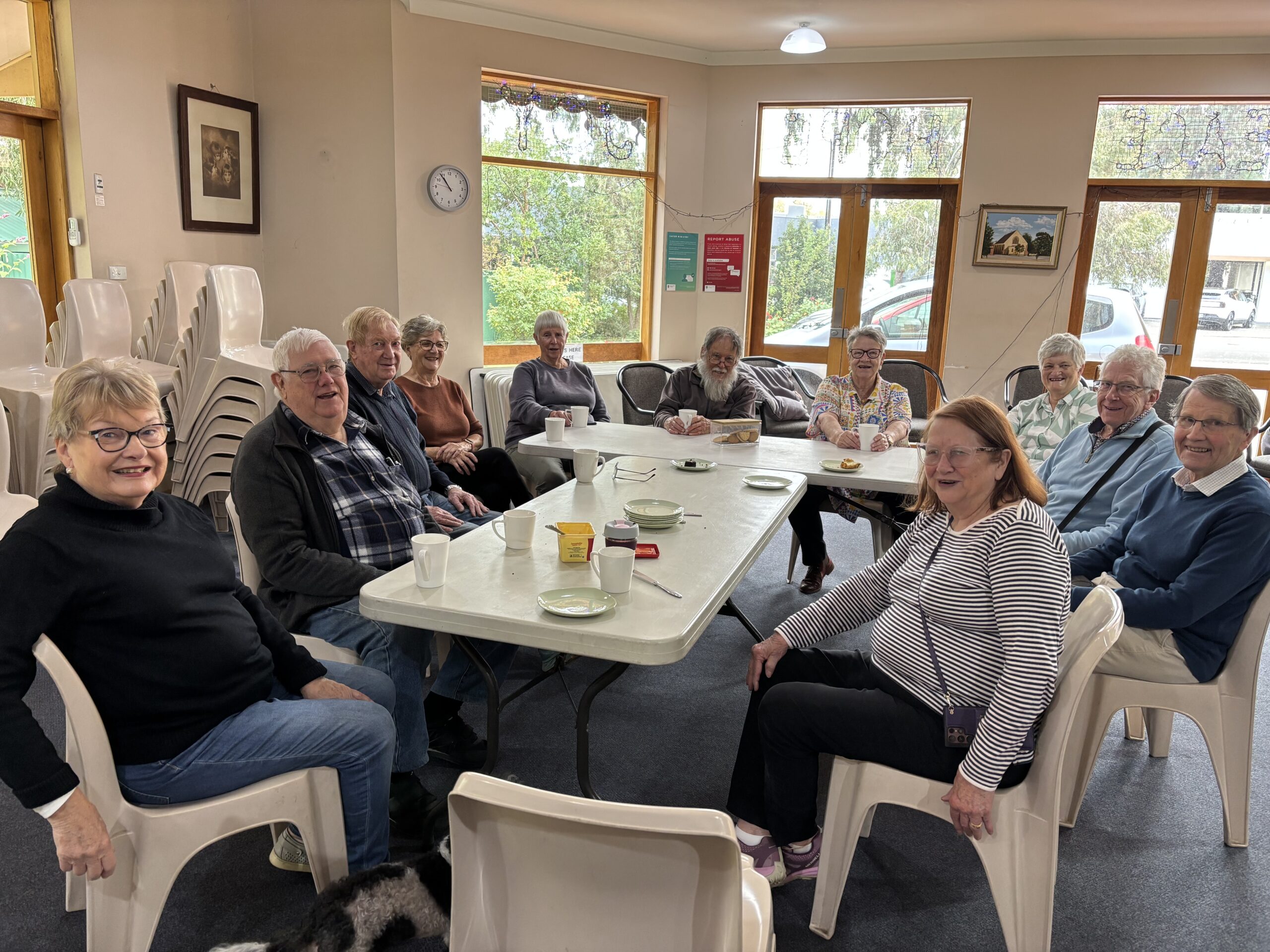 Group of people sitting around tables indoors with plates of biscuits and mugs.