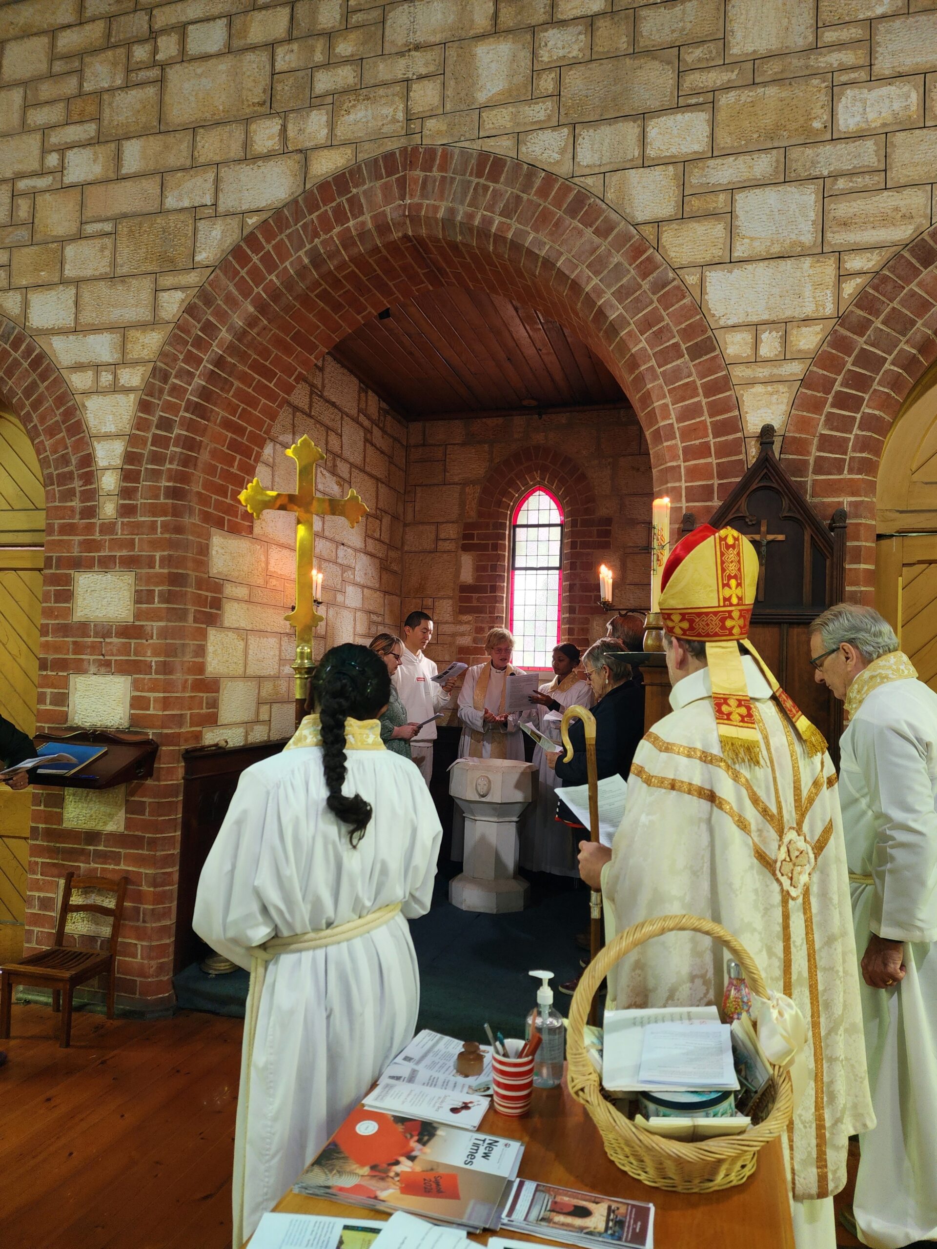 Priest, bishop, and group of laypeople standing around a font in an alcove at the back of a church.