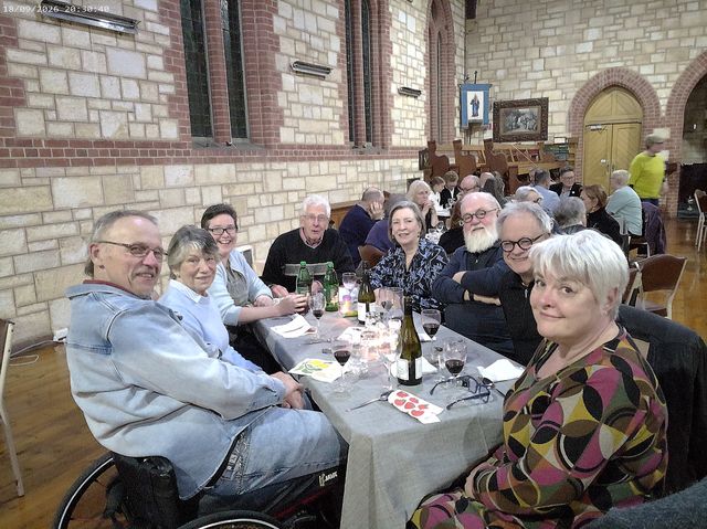 Group of smiling people sitting around a dining table with glasses and bottles in a church.